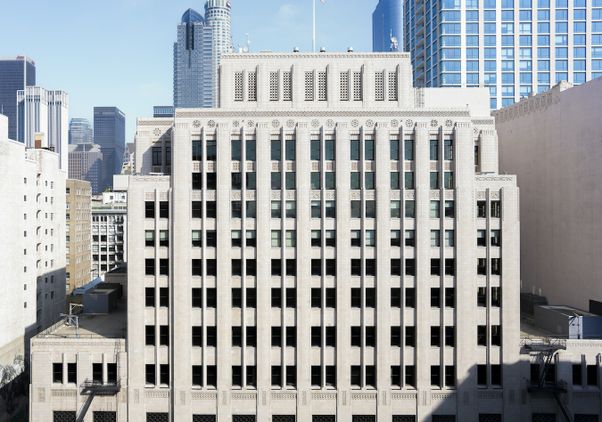 Exterior view of UCLA Downtown, a historic Art Deco building on Spring Street in downtown Los Angeles. The limestone façade features decorative architectural details, with modern skyscrapers rising behind the building beneath a clear blue sky.