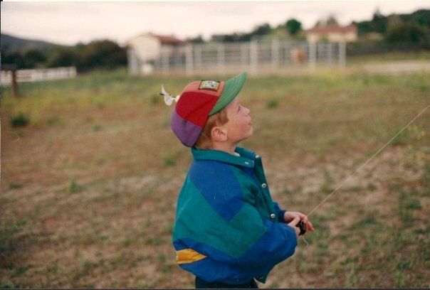 A childhood photo of Joey Curti as a young boy looking upward in an open field, wearing a colorful jacket and baseball cap.