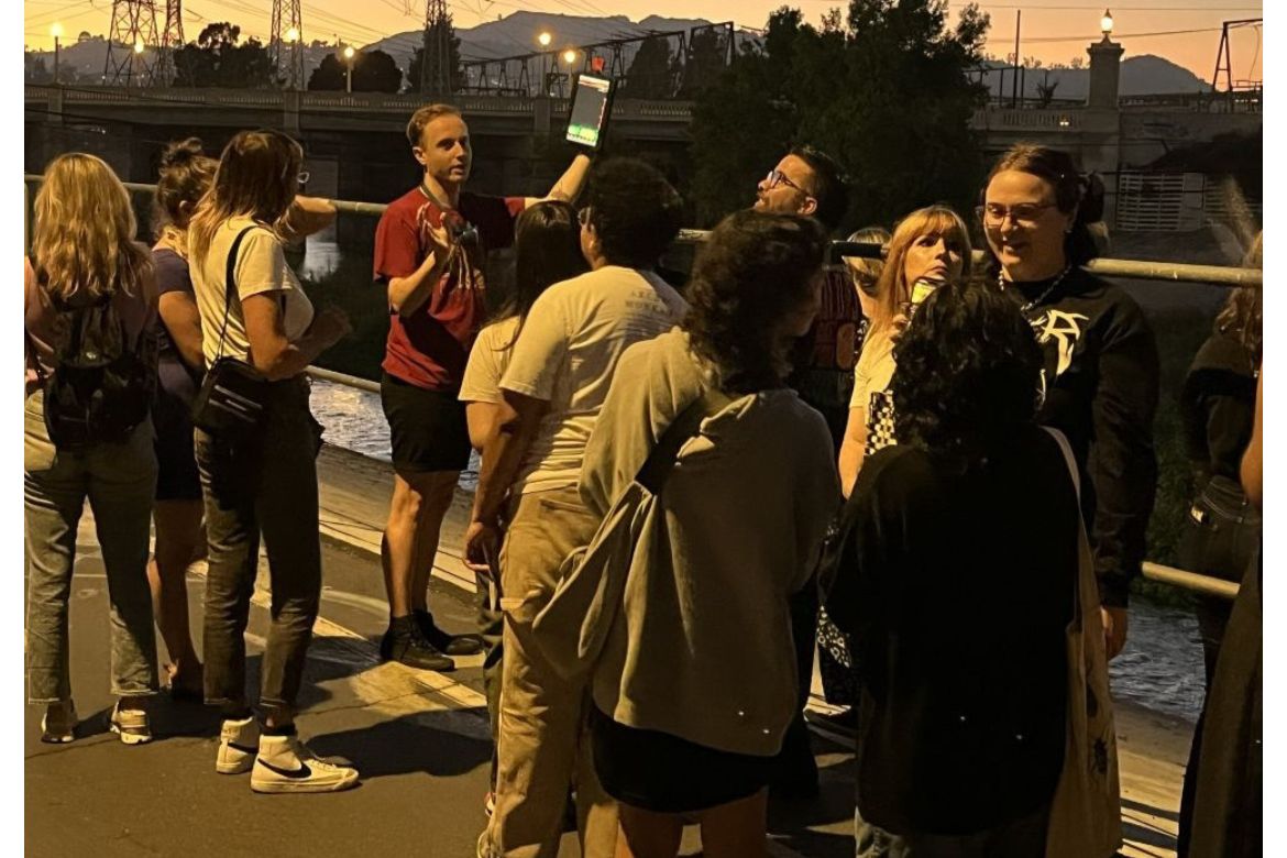 Joey Curti holds up a tablet while leading a group of people on a dusk bat walk along the Los Angeles River. A bridge and city lights are visible in the twilight background.
