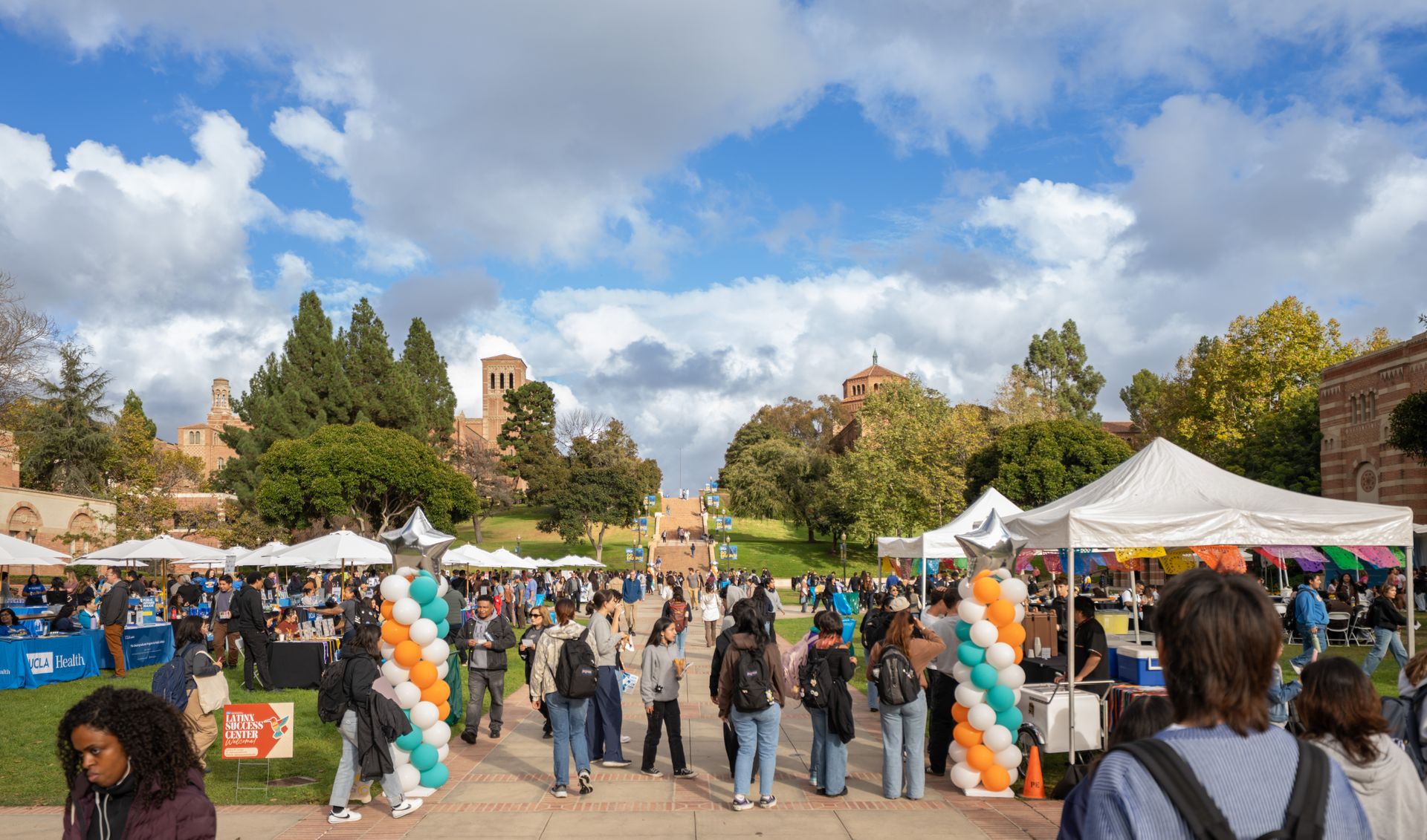 A wide view of Wilson Plaza at UCLA filled with students attending the Latinx Welcome event. White event tents, colorful balloon columns in teal, orange, and white, and UCLA campus buildings are visible under a partly cloudy sky.