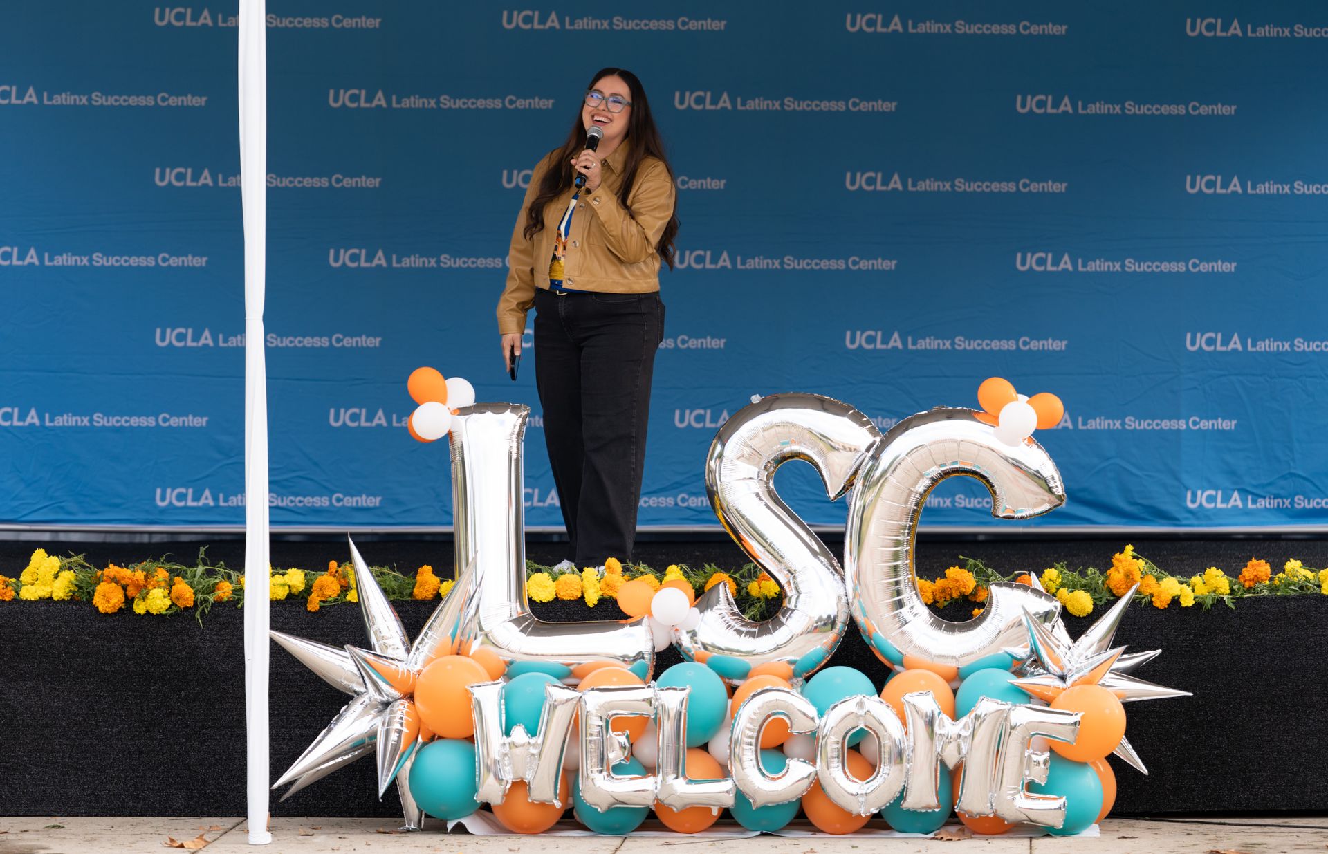 Arlene Cano Matute, executive director of the Latinx Success Center, speaks into a microphone on stage at the welcome event. In front of the stage, large silver balloon letters spell out "LSC Welcome," surrounded by teal, orange, and white balloons and marigold flowers.