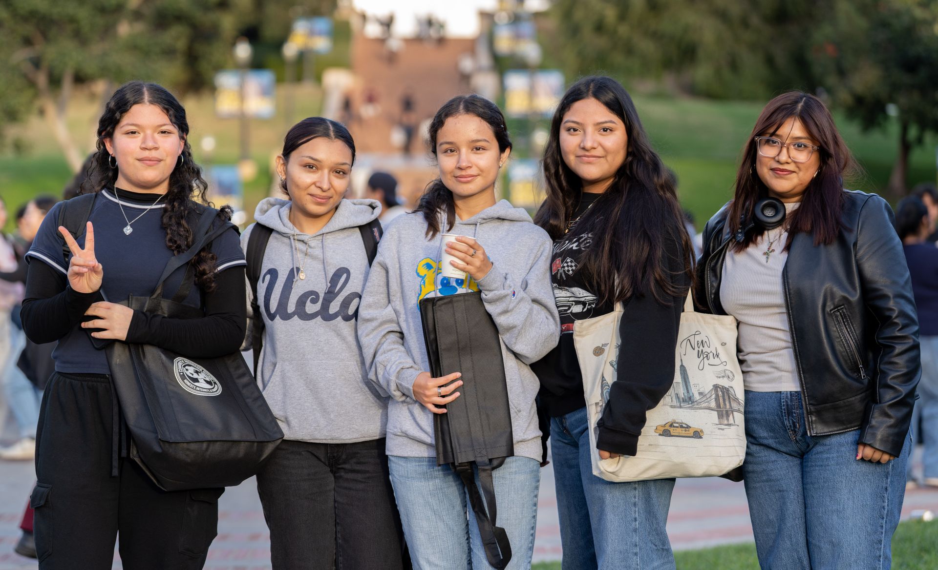 Five UCLA students smile and pose together on campus at the Latinx Success Center Welcome, holding giveaway items including tote bags and an umbrella. The UCLA campus walkway and buildings are visible in the background.