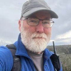 Outdoor selfie of Fred Ramsdell wearing glasses, a baseball cap, and a blue jacket, with a rocky hillside landscape in the background.