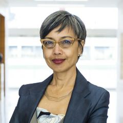 Headshot of Ninez A. Ponce, wearing glasses and a navy blazer, standing in front of a blurred indoor background.