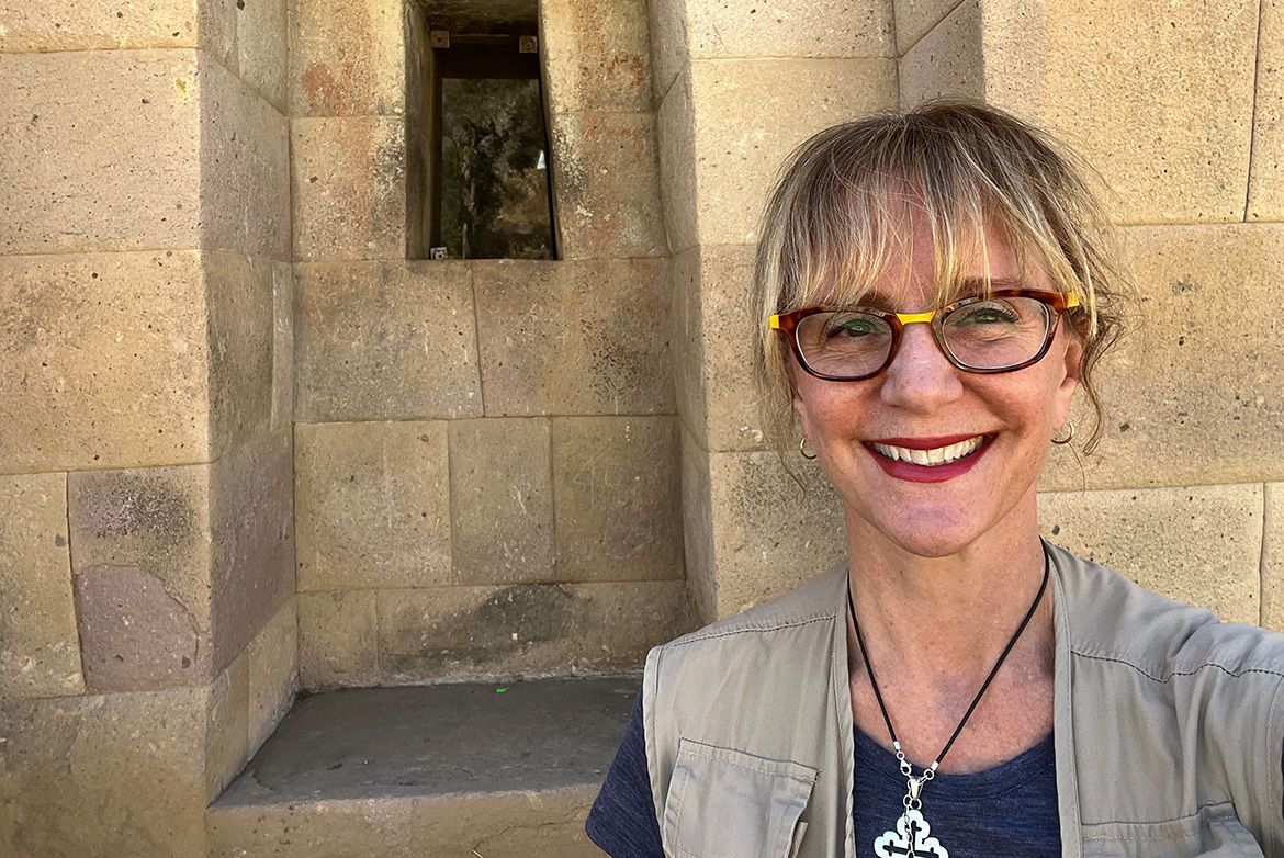 Stella Nair takes a selfie in front of an ancient stone wall with a trapezoidal Inca window niche at an archaeological site in Huaytará, Peru. She is smiling and wearing glasses, a gray vest, and a necklace.