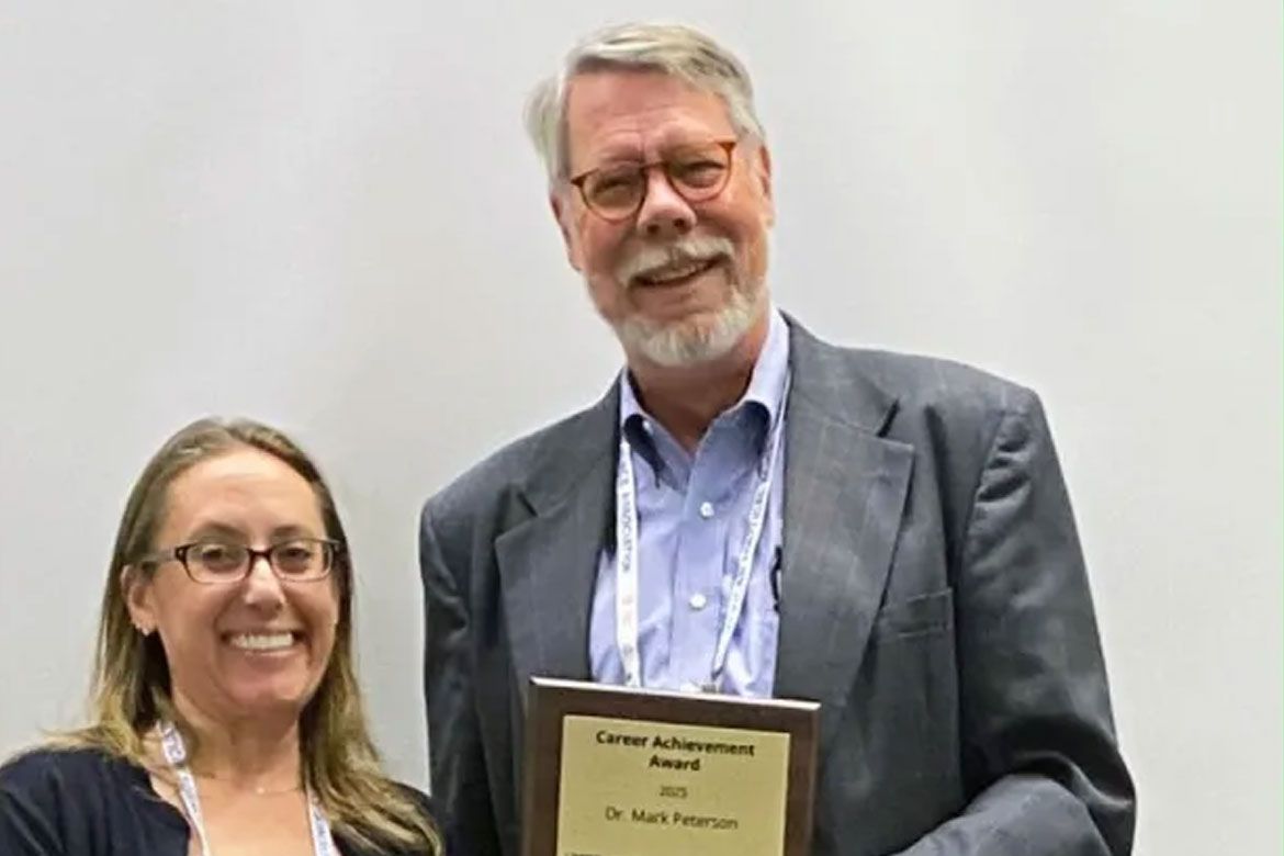 Julianna Pacheco and Mark Peterson smile while holding a plaque reading "Career Achievement Award, 2025, Dr. Mark Peterson." Both are wearing conference lanyards.