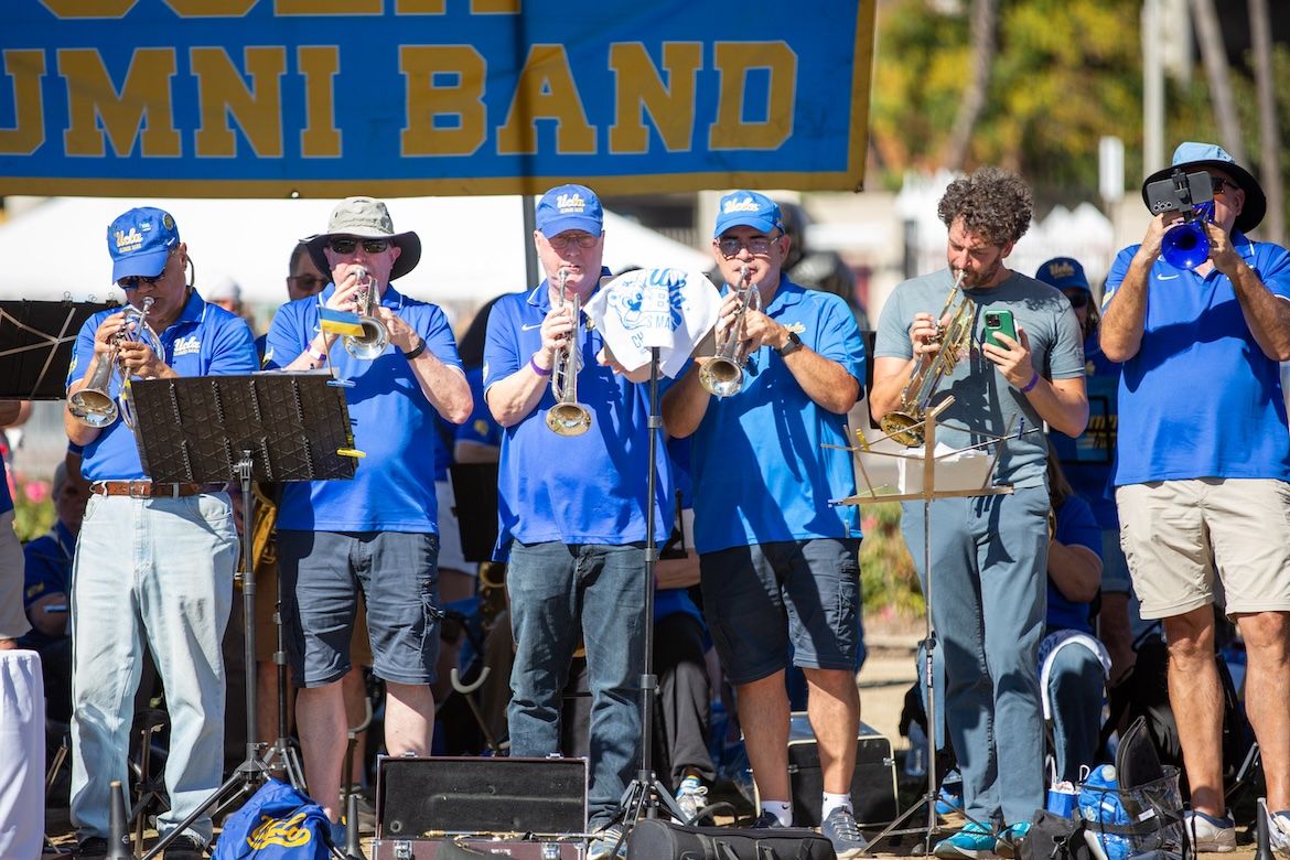 Members of the UCLA Alumni Band play trumpets on stage at a tailgate event. Most are wearing blue UCLA polo shirts and caps. A banner reading "UCLA Alumni Band" is visible above them.