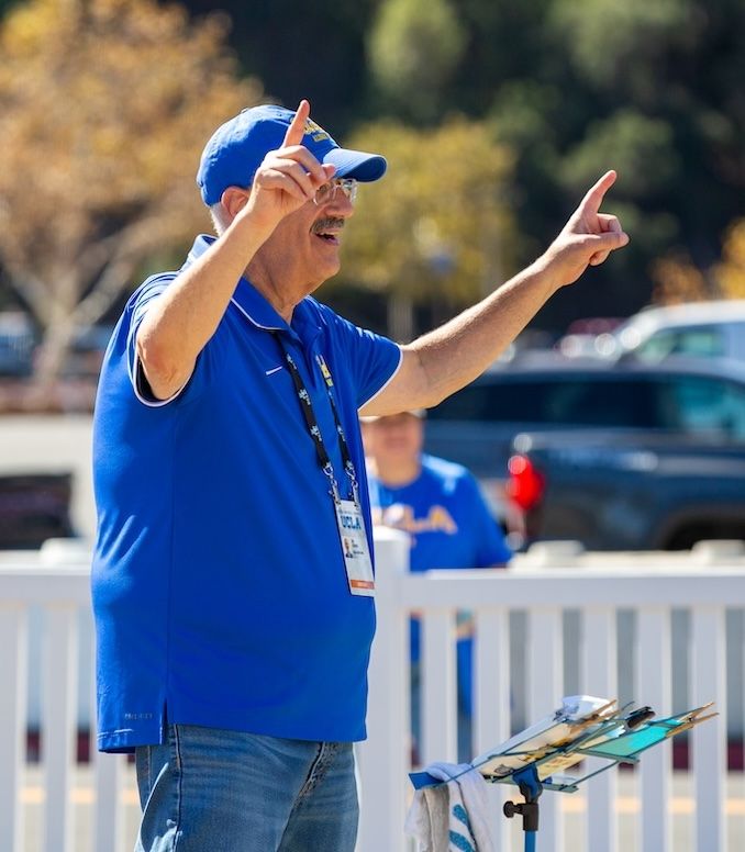Alumni band-Joel Fierberg Fierberg conducts the UCLA Alumni Band at the October 18 Rose Bowl tailgate, pointing both index fingers upward while wearing a blue UCLA polo shirt and cap. A music stand is visible in the foreground.
