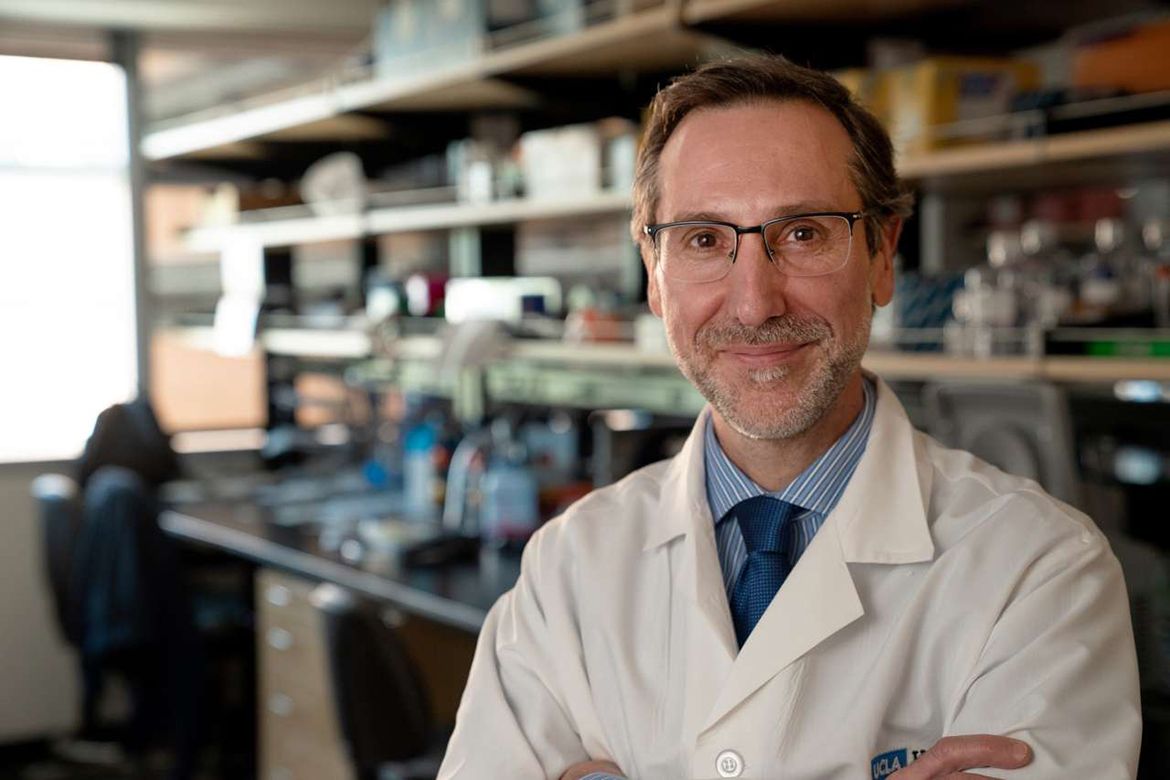 Dr. Antoni Ribas, wearing a white lab coat and blue tie, stands with arms crossed and smiles at the camera in a research laboratory. 