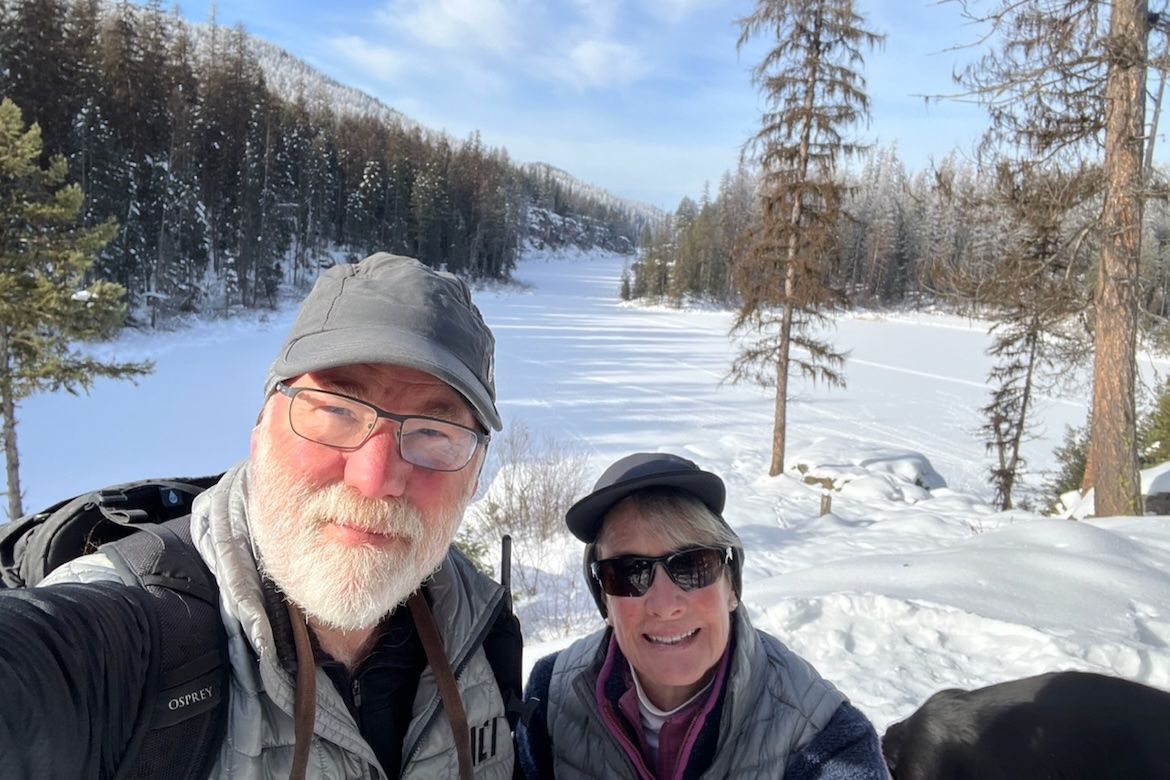 Ramsdell and O'Neill take a selfie on a snow-covered trail surrounded by snow-dusted evergreen trees and a frozen landscape under a blue sky. Both are wearing winter gear including hats, sunglasses, and vests. 