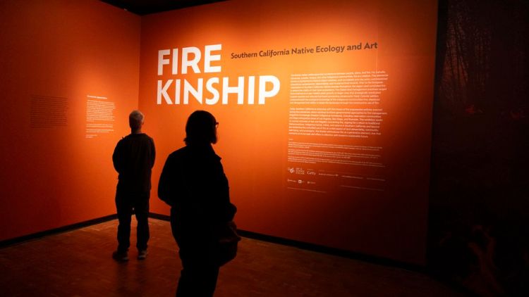 Two visitors stand silhouetted before a glowing orange entrance wall reading "Fire Kinship: Southern California Native Ecology and Art" at a museum exhibition.
