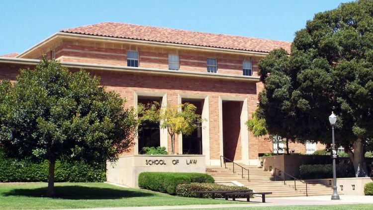 Exterior of the UCLA School of Law building, a red brick structure with large columns and steps leading to the entrance, surrounded by trees and a green lawn under a clear blue sky.