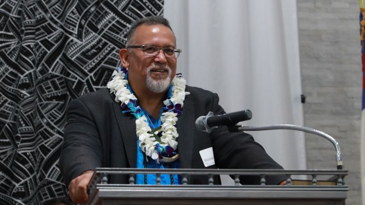 Vicente Diaz wearing glasses, a blue shirt, and a white and blue floral lei speaks and smiles at a podium in front of a black and white patterned textile backdrop.