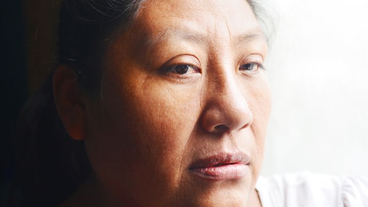 Close-up portrait of a woman with dark hair pulled back, looking thoughtfully off to the side, with soft natural light illuminating her face.