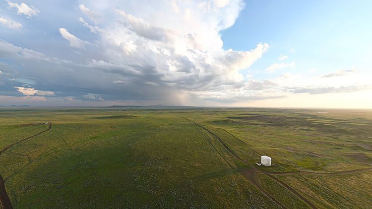 Aerial view of a vast, flat green prairie (Fort Belknap Indian Reservation) under a dramatic sky with large storm clouds and a distant rain shower, with a dirt road and a small white structure visible below.