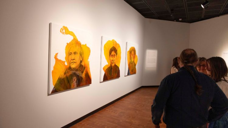 Museum visitors view three large paintings on a white gallery wall depicting portrait figures with warm golden and orange backgrounds, in a dimly lit exhibition space.