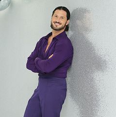 Valentin (Val) Chmerkovskiy, professional dancer on Dancing With the Stars, leans against a textured gray wall with arms crossed, wearing a purple long-sleeve shirt and matching pants.
