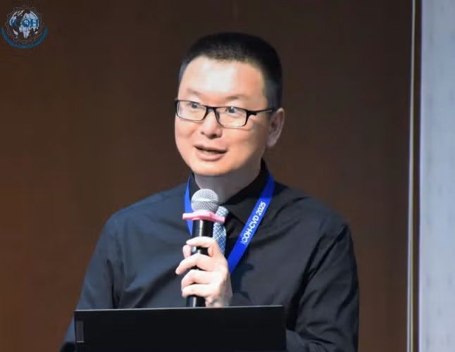 Jian Li speaks at a podium, holding a microphone. He is wearing glasses, a dark button-down shirt, and a blue conference lanyard.