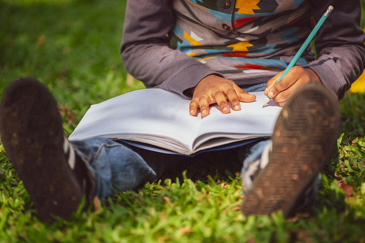 A child, seen from the shoulders down, sits on grass with their legs out and a book on their lap