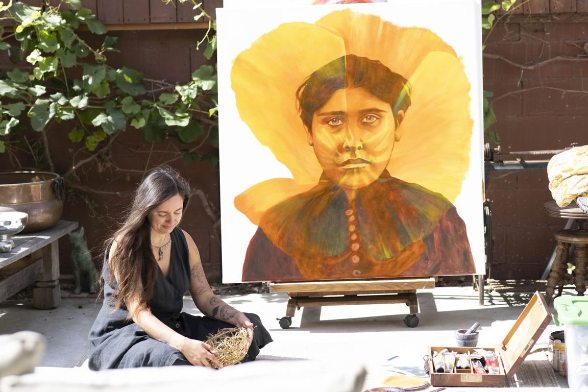 Artist Weshoyot Alvitre sits beside her large portrait painting "California Native Dormidera #2: Modesta Avila" on an outdoor easel, holding a woven nest, with paint supplies nearby.