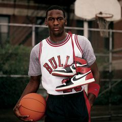 A young Michael Jordan stands on an outdoor basketball court in the 1980s, wearing a Chicago Bulls uniform and holding a basketball under one arm and a pair of original Air Jordan 1 sneakers in his other hand.