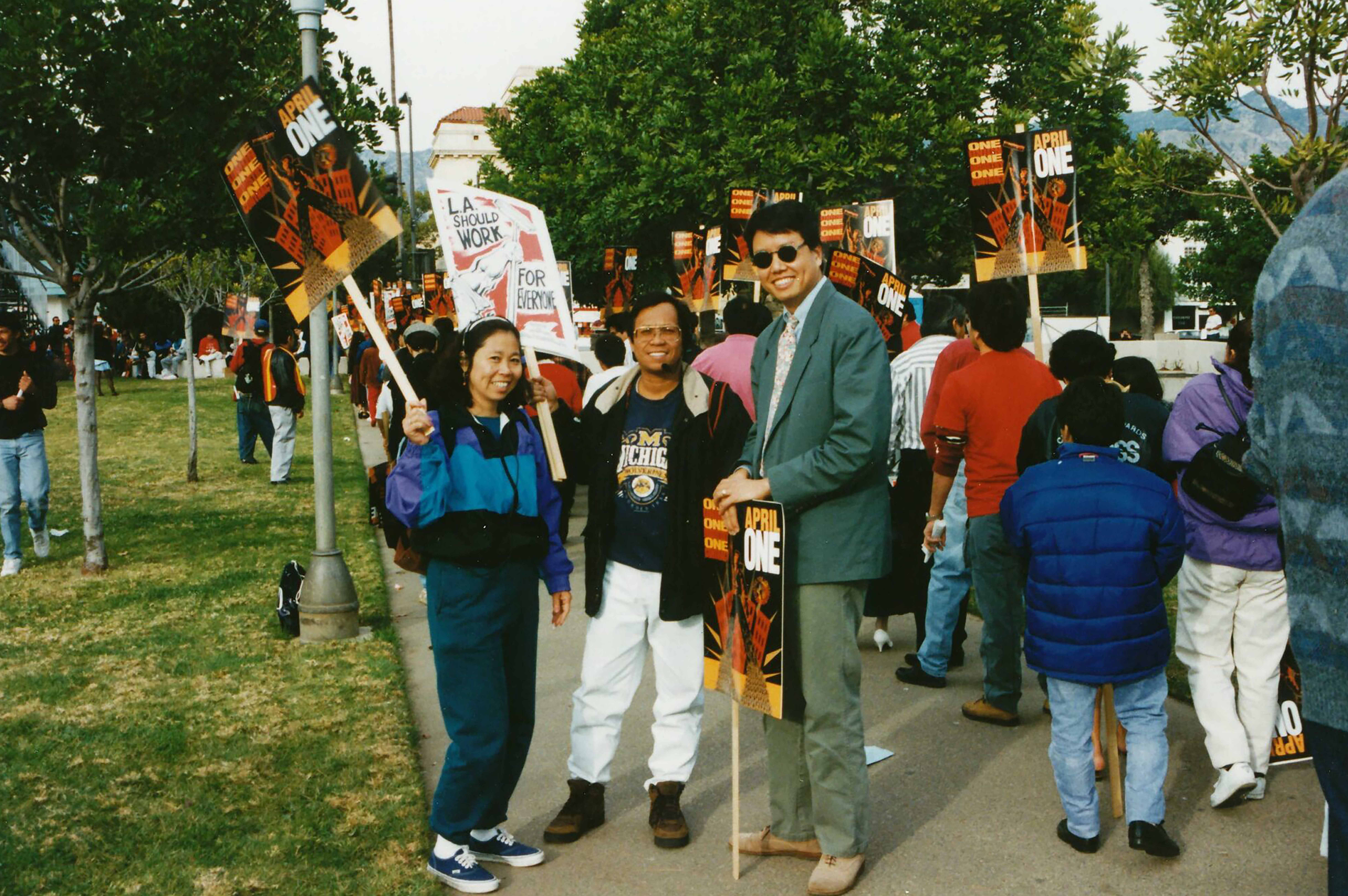 Kent Wong, right, in gray jacket and sunglasses, leans on a sign as he stands next to a woman and man who are holding up signs
