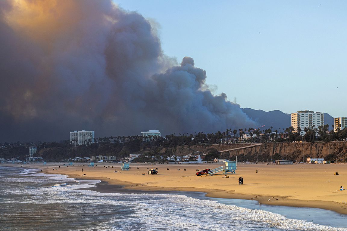 A massive plume of smoke rises from the Palisades fire on Jan. 7, 2025, visible from the beach with coastal buildings and cliffs in the foreground.