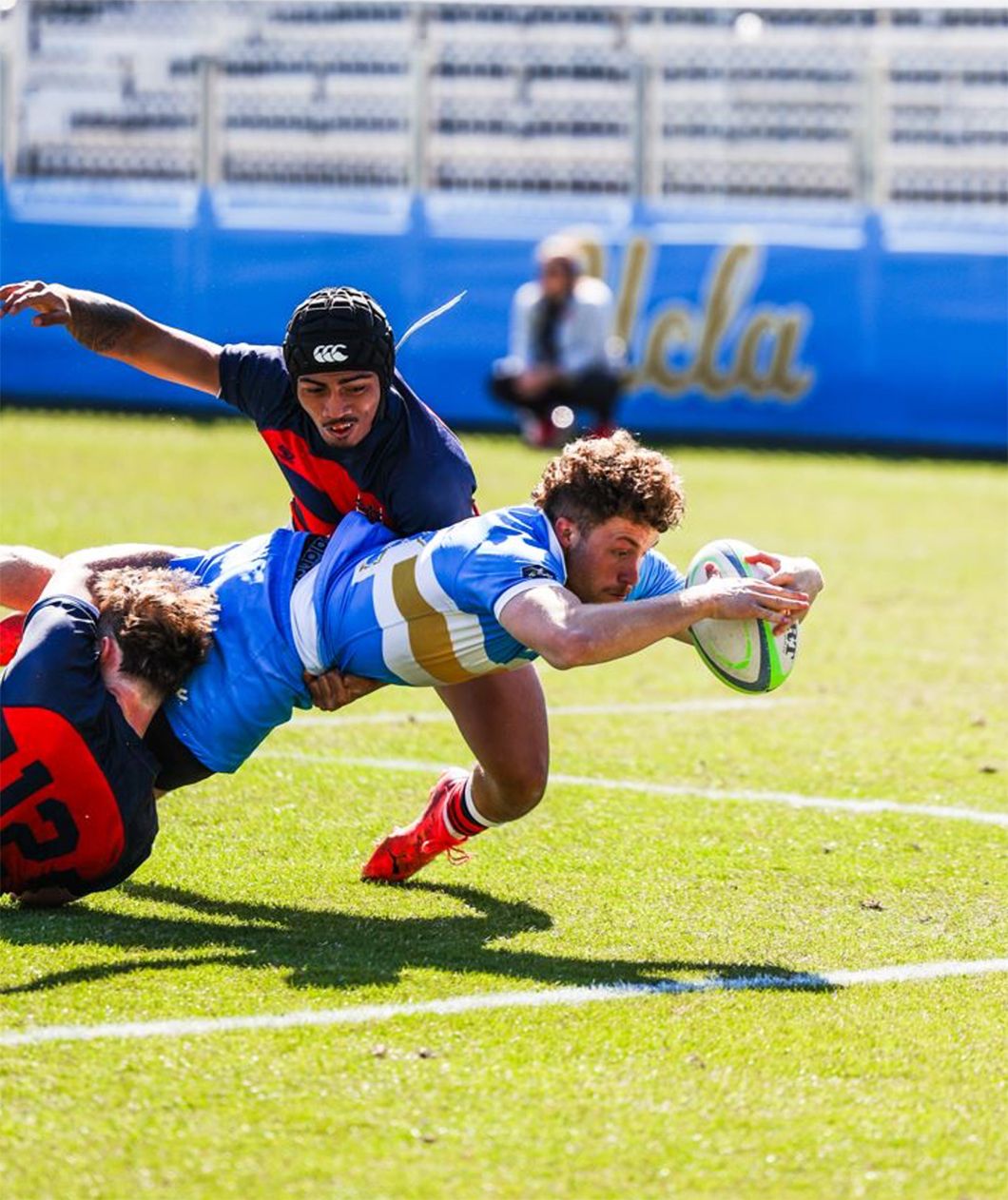Rugby player in blue and gold getting tackled by two players in navy and red