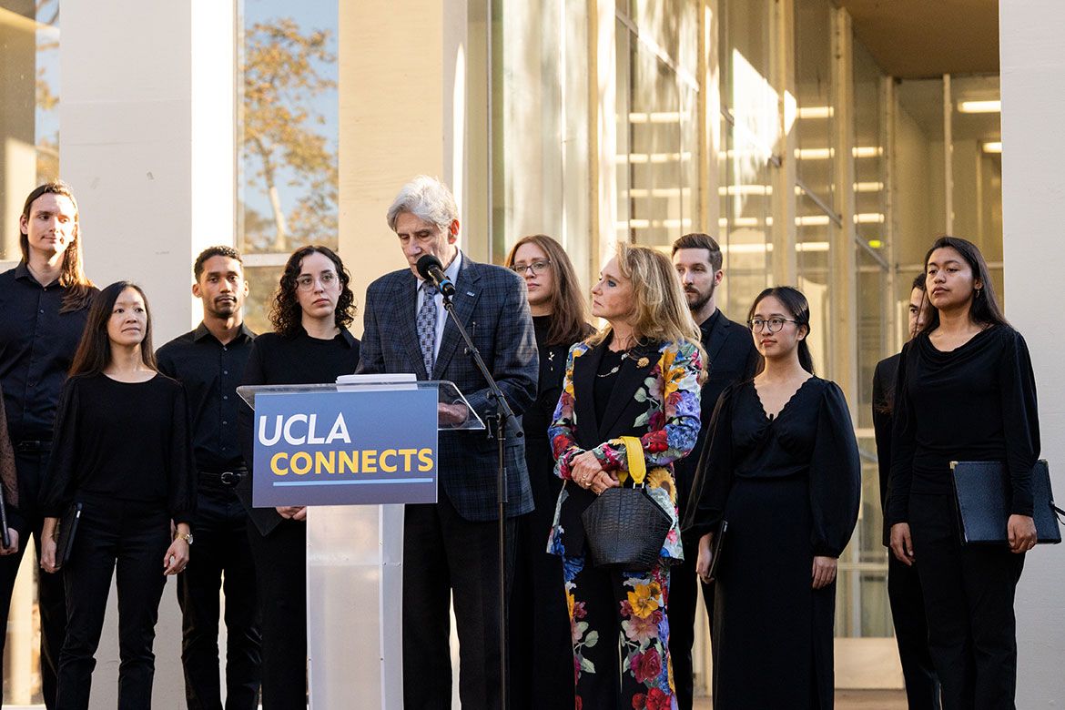 UCLA Chancellor Julio Frenk speaks at a UCLA Connects podium outdoors, flanked by Felicia Marie Knaul and a group of students dressed in black.
