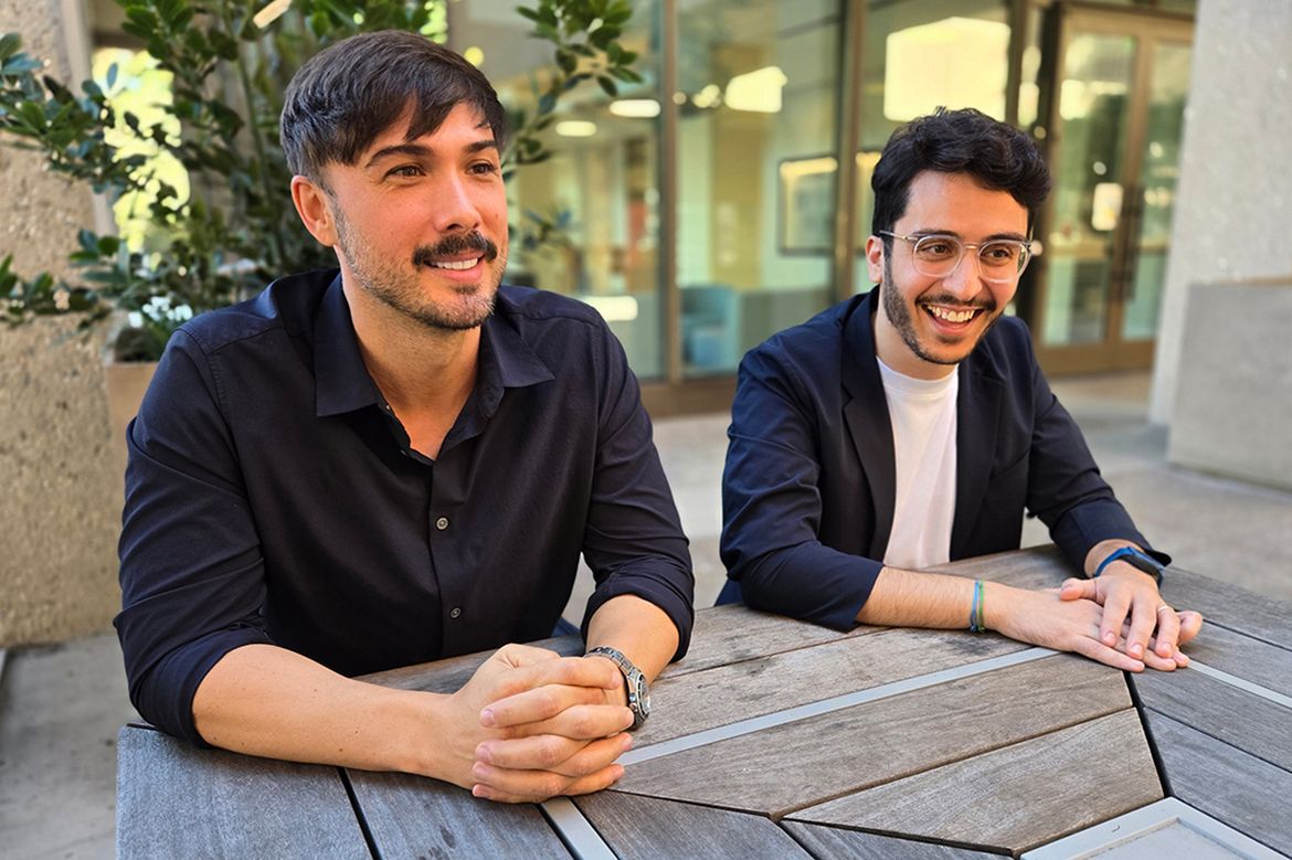 Richard Assaley (left), in dark blue button-down, and Glen Spiteri, in dark jacket and white T-shirt, sit at table with hands clasped