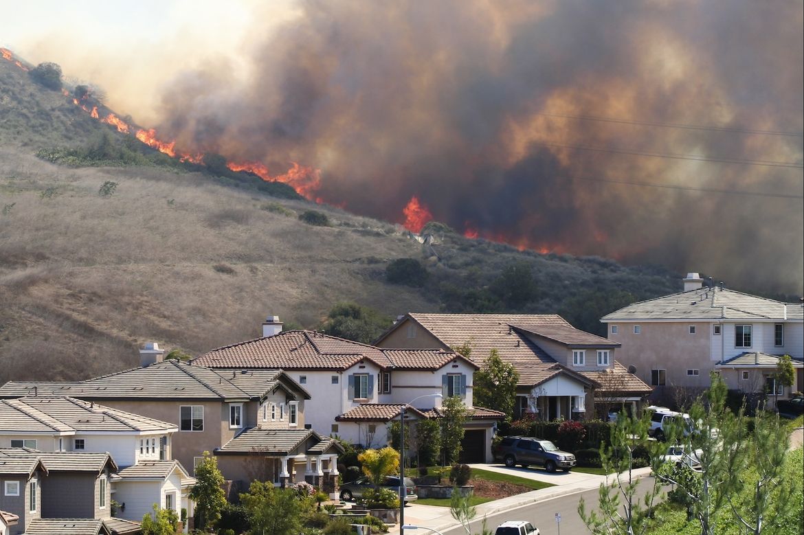 A wildfire burns along a steep hillside directly above a suburban neighborhood, with flames and heavy smoke visible over closely spaced residential homes.