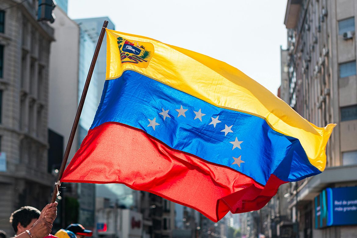 A person waves a Venezuelan flag with yellow, blue, and red stripes and seven white stars on a city street.
