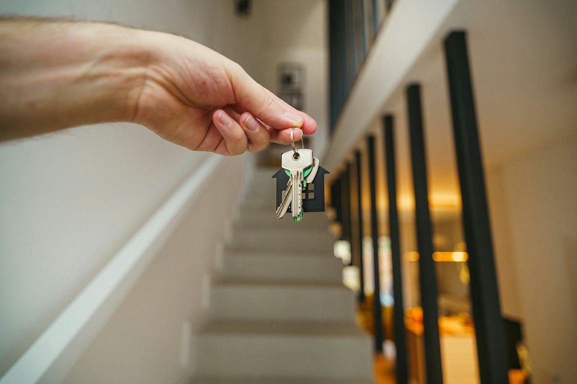A hand holds a set of house keys with a house-shaped keychain in the foreground of a residential staircase.