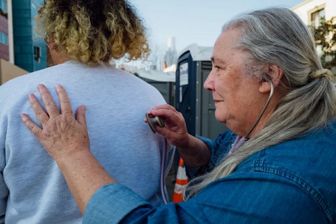 A health care provider with long gray hair uses a stethoscope to examine a patient's back at an outdoor medical clinic.