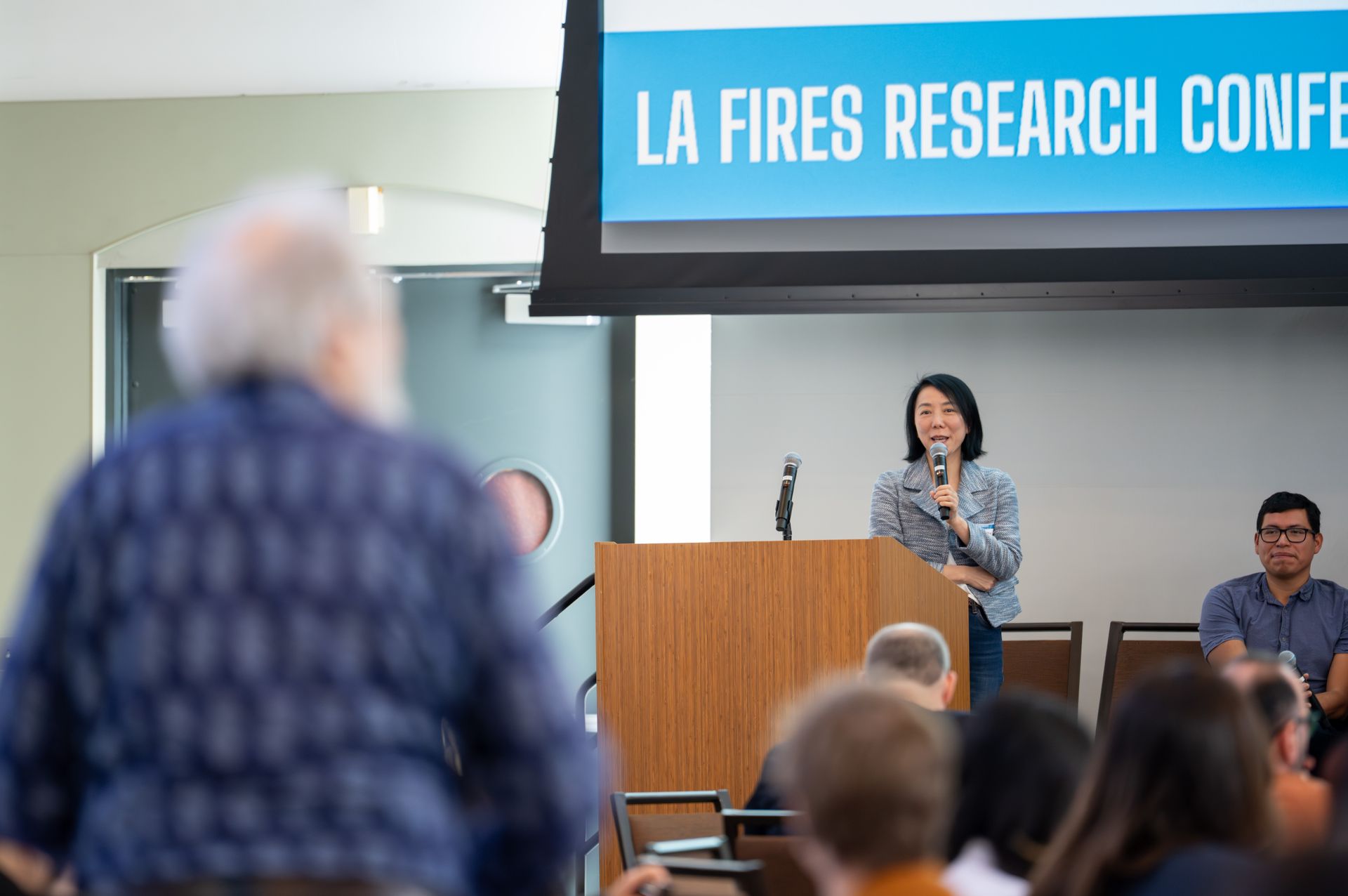 UCLA's Yifang Zhu speaks at a podium beneath a screen reading 'LA Fires Research Conference,' addressing audience members about communicating wildfire health risks to the public.