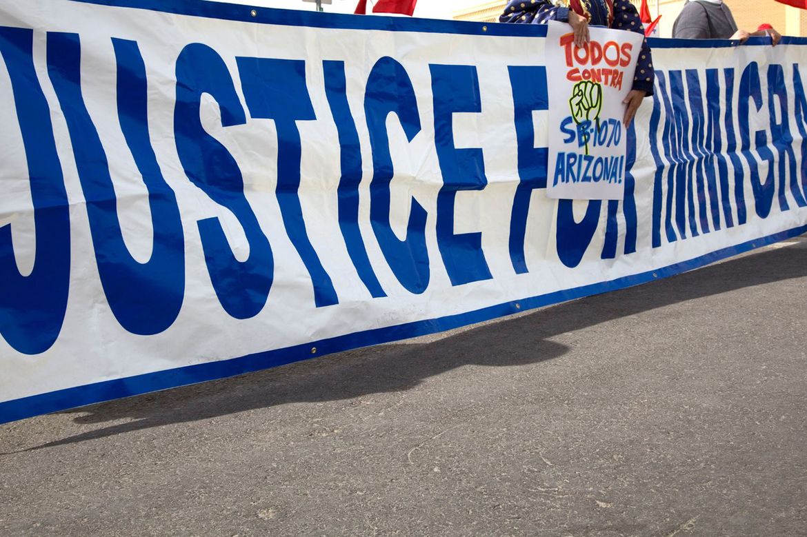 A large blue and white banner reading 'Justice for Immigrants' at an outdoor rally, with a smaller sign visible reading 'Todos Contra SB 1070 Arizona.