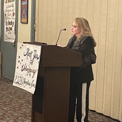 Felicia Marie Knaul speaks at a wood podium in front of a handwritten thank-you sign.