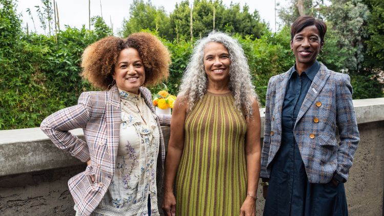 Dean Tracy Johnson in a floral blazer, Professor Gina Poe in a green sleeveless dress with long gray hair, and Bruin alumna Bridgid Coulter Cheadle in a plaid blazer, posing together outdoors against a lush green backdrop at a panel discussion on diversity.