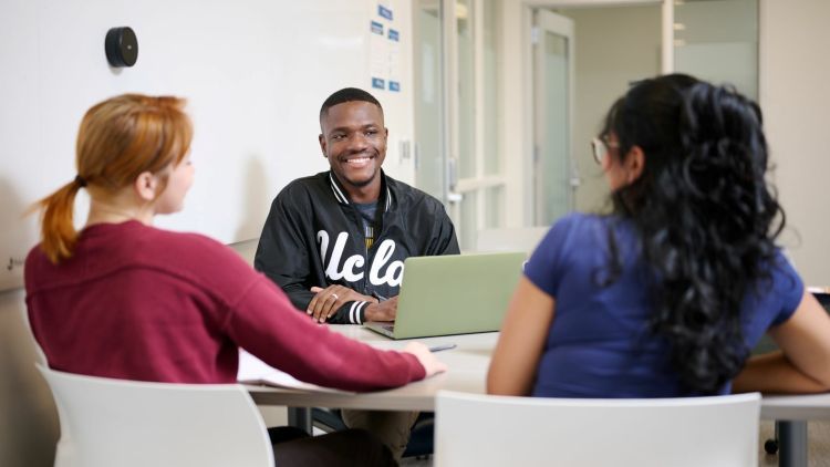 Prince Larmie Jarbo, wearing a UCLA jacket, smiling while seated with two fellow students at a campus table with a laptop.