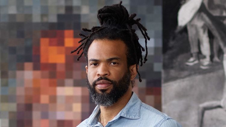 A portrait of Cosmo Whyte, UCLA School of the Arts and Architecture assistant professor, wearing a denim shirt with his hair in locs, standing before two artworks: a colorful pixelated painting on the left and a black-and-white figurative photograph on the right.