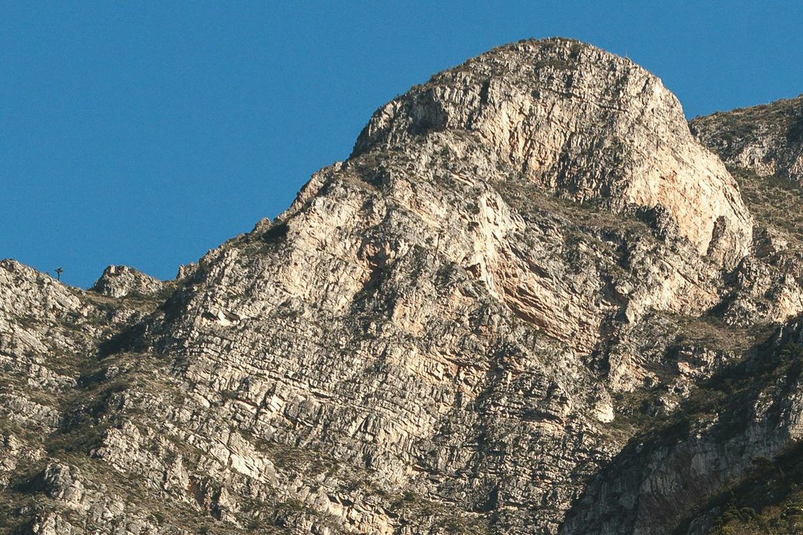 A rugged, rocky mountain face with layered limestone cliffs and sparse vegetation under a clear blue sky. Sunlight illuminates the upper peaks, highlighting the textured, fractured rock surface typical of the Andes mountain range.