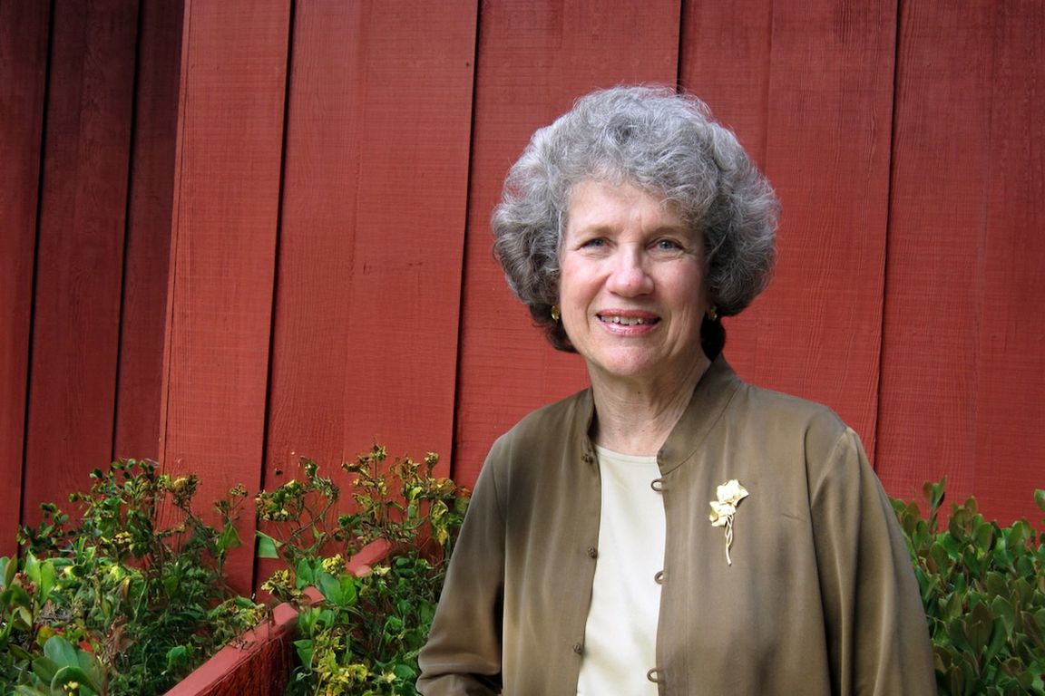 A portrait of Aimee Dorr smiling outdoors, wearing a brown jacket with a floral brooch, in front of a red wooden wall and greenery.