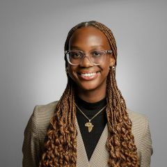 Headshot of Laila Wheeler, smiling, wearing a blazer and necklace with an Africa-shaped pendant, with long braided hair.