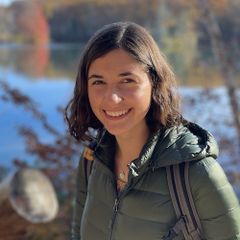 Headshot of Laurel Thomas, smiling, wearing a green jacket, photographed outdoors near autumn foliage and water.