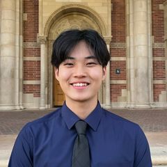 Headshot of Michael Suh, smiling, wearing a dark blue shirt and tie, with a brick building visible in the background.