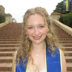 Headshot of Emma Montilla, smiling, with long curly blonde hair, wearing a blue graduation stole on steps outdoors.