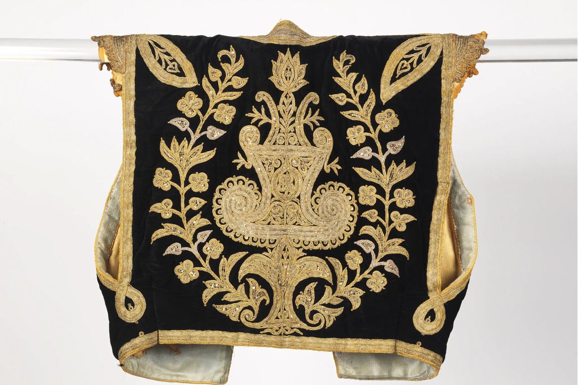 A historic black velvet garment heavily embroidered with intricate gold floral and botanical designs, displayed on a hanger against a white background.