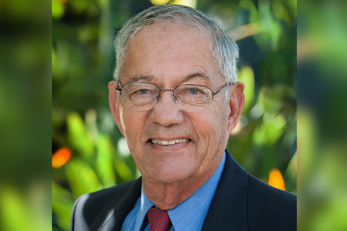 A portrait of Morton La Kretz smiling outdoors, wearing glasses, a dark blazer, and a red tie.