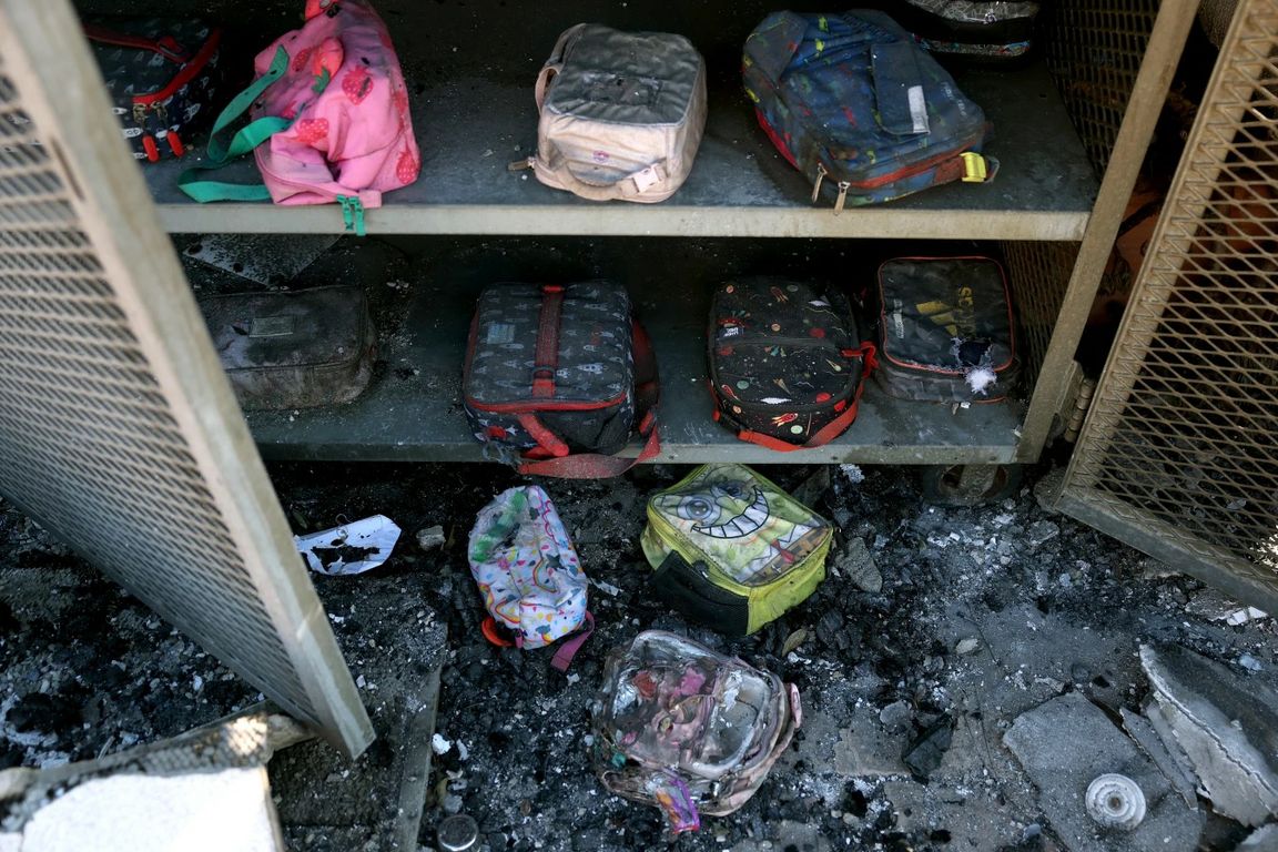 Children's backpacks and lunch boxes on charred shelves surrounded by ash and debris in the aftermath of a fire."