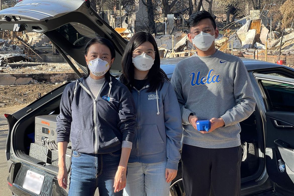 Professor Yifang Zhu, researcher Yuan Yao, and postdoctoral scholar Haoxuan Chen, all wearing respirator masks, stand beside a car with research equipment amid wildfire destruction in Los Angeles County.